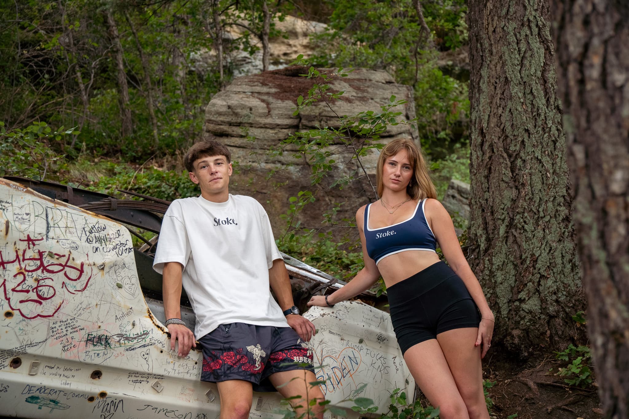 Two people outside in Stoke — the white Everything Tee and the navy Everything Bra — leaning on a graffiti-covered wrecked car in the woods